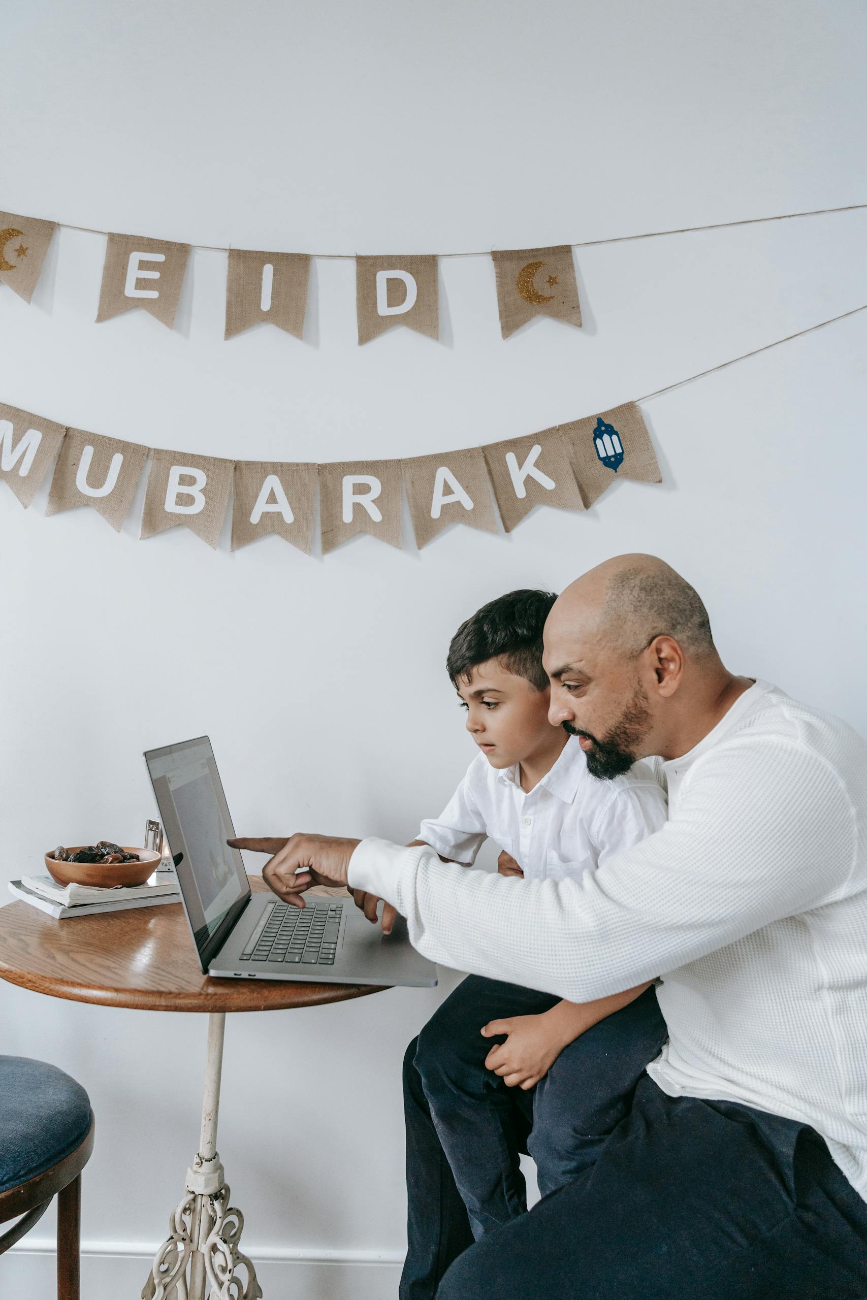 Father and son engaging with laptop during Eid celebration, enjoying quality time indoors.